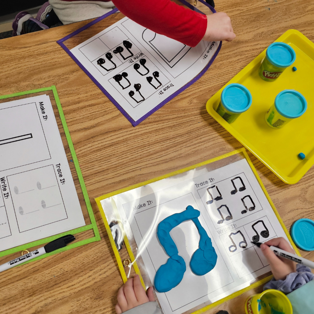 a yellow tray has three blue play doh containers on it. There are also two children with music themed dry erase mats drawing music notes .Their mats have play dough notes on them. 